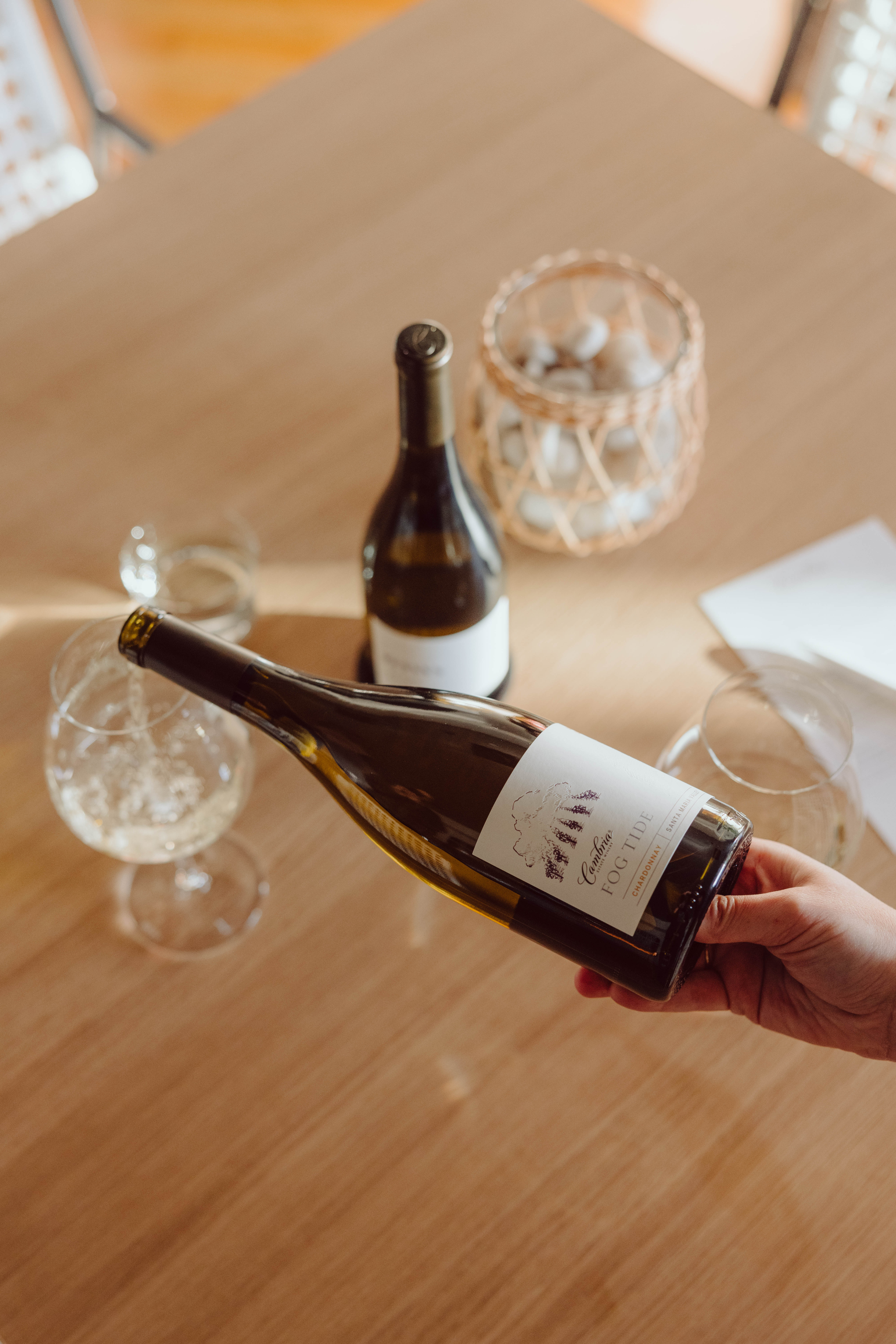 A hand pouring a white wine into a wine glass with a bottle and glass on the table beneath.